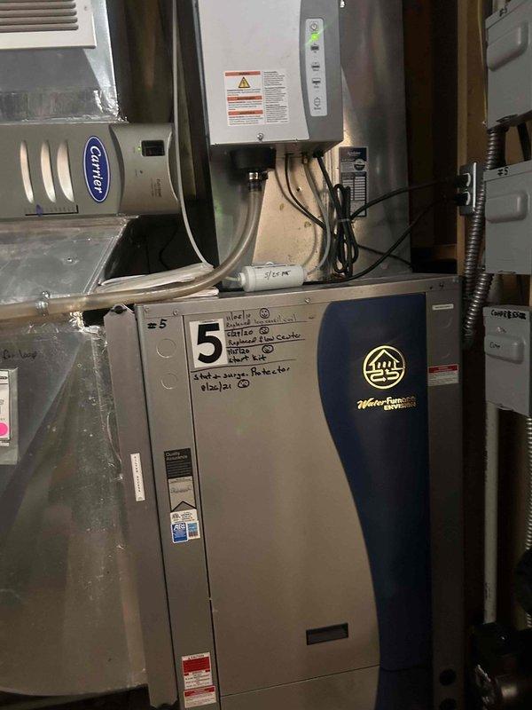 Responded to no cooling call for residential property with multiple geothermal heat pump systems. Upon arrival, found customer operates 7 WaterFurnace Envision geothermal units throughout the home. Conducted systematic inspection of units, focusing on unit #5 and unit #7 as photographed during diagnostic process. Evaluated electrical connections, control boards, and thermostat operations on affected systems. Inspected wall-mounted controls showing indicator lights and verified proper communication between thermostats and air handlers. Checked Carrier-branded air handling components and associated ductwork for airflow restrictions. Reviewed service history notations marked on equipment cabinets dating back to previous maintenance visits. Assessed geothermal loop connections and blue buffer tanks attached to units. Determined which of the 7 geothermal systems were experiencing cooling failures versus operating normally. Documented equipment conditions with photographs for service records