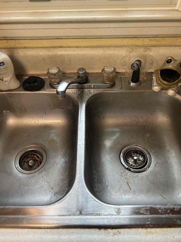Responded to service call for kitchen faucet leak with unknown location. Upon arrival, performed comprehensive inspection of double-basin kitchen sink system with dual-handle chrome faucet assembly. Identified source of leak at corroded horizontal copper supply line connection beneath sink. Supply lines exhibited significant corrosion, oxidation, and green patina buildup indicating long-term deterioration. Shut off water supply via angle stop valves and assessed condition of all under-sink plumbing components. Documented extensive mineral deposits throughout sink basins, aged drain assemblies, and compromised braided flexible supply lines. Recommended replacement of corroded copper piping, supply lines, and shut-off valves to prevent future leaks and ensure reliable water delivery to faucet system.