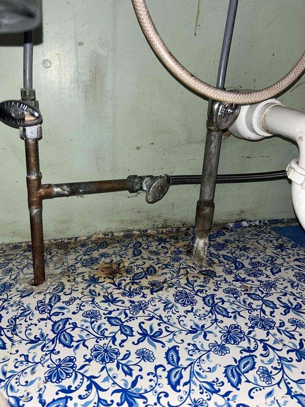 Responded to service call for kitchen faucet leak with unknown location. Upon arrival, performed comprehensive inspection of double-basin kitchen sink system with dual-handle chrome faucet assembly. Identified source of leak at corroded horizontal copper supply line connection beneath sink. Supply lines exhibited significant corrosion, oxidation, and green patina buildup indicating long-term deterioration. Shut off water supply via angle stop valves and assessed condition of all under-sink plumbing components. Documented extensive mineral deposits throughout sink basins, aged drain assemblies, and compromised braided flexible supply lines. Recommended replacement of corroded copper piping, supply lines, and shut-off valves to prevent future leaks and ensure reliable water delivery to faucet system.