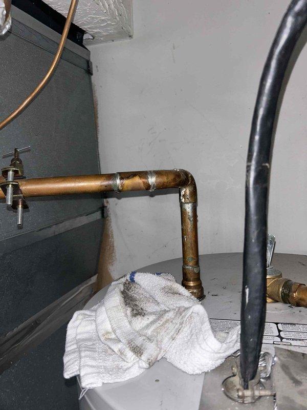 Responded to service call for 12-year-old tanked water heater in garage with no hot water and active leaking. Upon inspection, identified significant leak at upper pipe connections with visible water damage and towels placed to absorb moisture around copper piping. Assessed unit condition, noting age-related deterioration including oxidation and patina on copper lines and brass fittings. Documented equipment configuration including connections to electrical panel, multiple pipe runs (PVC and copper), and overall system layout. Photographed leak points and surrounding plumbing infrastructure for diagnostic evaluation and replacement recommendations given unit age and extent of failure.
