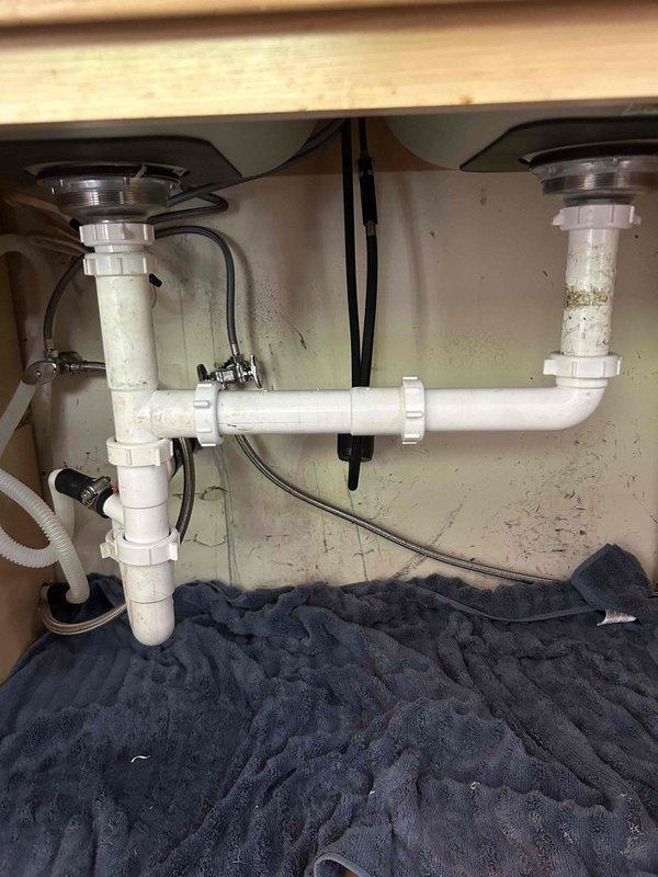 Responded to callback for leak under recently replaced kitchen sink. Upon inspection, identified active leak at drain assembly connection. Disassembled P-trap configuration and drain connections, finding improper seal at basket strainer. Reinstalled drain components with proper sealing, ensuring all PVC connections were secure and watertight. Tested system with running water to confirm leak was resolved. Photographed completed work showing proper P-trap installation and cabinet interior condition. Removed old fittings and materials from site.