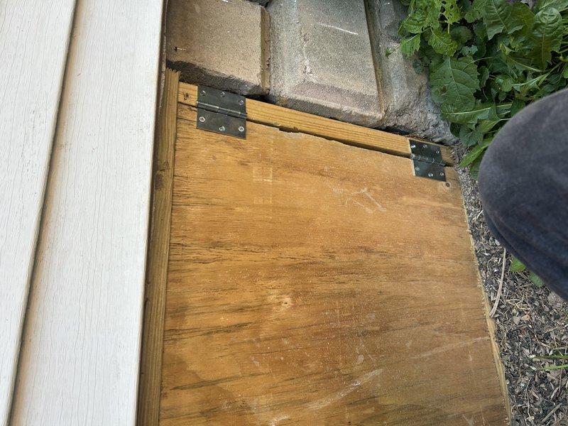 Completed installation of wooden access door with black metal hinges to cover HVAC system at exterior crawlspace entrance. Door was secured to concrete block foundation adjacent to vinyl siding, with latch mechanism installed for proper closure. Photographic documentation taken to verify completion of work that was originally scheduled as part of initial system installation.