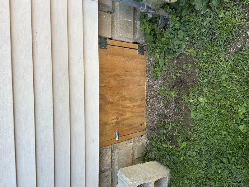 Completed installation of wooden access door with black metal hinges to cover HVAC system at exterior crawlspace entrance. Door was secured to concrete block foundation adjacent to vinyl siding, with latch mechanism installed for proper closure. Photographic documentation taken to verify completion of work that was originally scheduled as part of initial system installation.