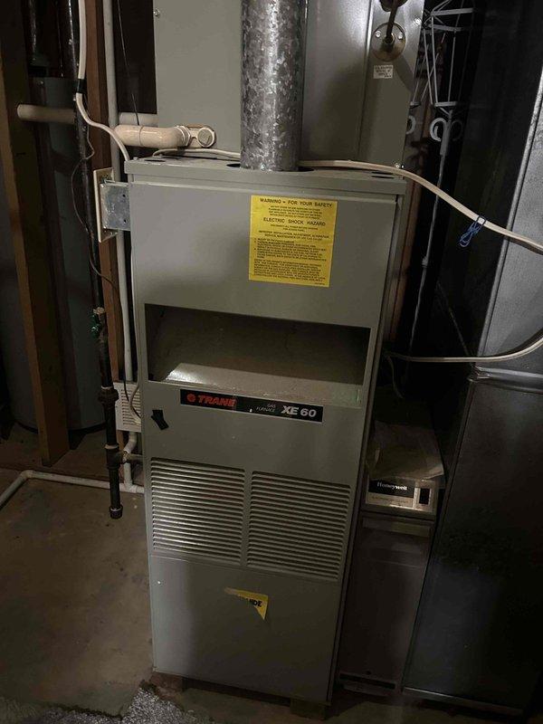 Responded to no cooling complaint on 6-year-old gas furnace/AC system with outdoor unit not running and indoor unit blowing ambient temperature air. Upon arrival, performed diagnostic evaluation of the system including electrical testing of the outdoor condensing unit. Used clamp meter to verify power supply and measure electrical parameters at the condenser. Inspected Trane XE60 gas furnace and associated ductwork for proper operation and airflow. Identified issue preventing outdoor unit operation and documented equipment conditions with diagnostic photos for service records.