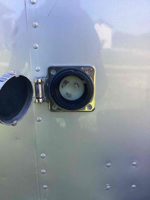 Completed electrical estimate for powering an Airstream trailer. Assessed existing twist-lock style power inlet receptacle mounted on the trailer's exterior aluminum panel and evaluated the yellow weatherproof connector outlet positioned at ground level near the unit. Determined electrical requirements and connection specifications for establishing proper 120/240V service to the recreational vehicle.