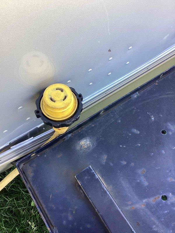 Completed electrical estimate for powering an Airstream trailer. Assessed existing twist-lock style power inlet receptacle mounted on the trailer's exterior aluminum panel and evaluated the yellow weatherproof connector outlet positioned at ground level near the unit. Determined electrical requirements and connection specifications for establishing proper 120/240V service to the recreational vehicle.