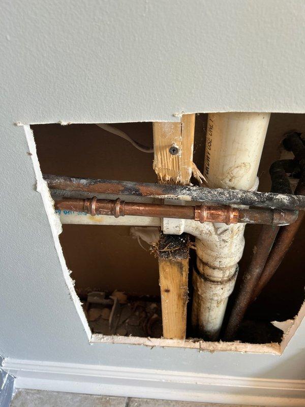 Responded to a pipe leak in a 20+ year old residential property. Upon inspection, accessed the affected plumbing through ceiling/wall cavity and identified a compromised copper water line showing significant age-related corrosion and oxidation. Removed drywall to expose the plumbing chase, revealing aged copper piping with darkened patina and PVC drain/vent lines with weathering and discoloration. Assessed the leak source at soldered joint connections where debris accumulation and pipe deterioration were evident. Installed ball valve isolation point on the copper supply line to control water flow to the affected section. Verified proper operation of shutoff valve and confirmed leak containment. Inspected surrounding wooden framing and adjacent green PVC piping for secondary water damage or structural concerns. The plumbing system displayed typical wear consistent with the home's age, with multiple material transitions between copper supply lines and plastic drainage components. Access 