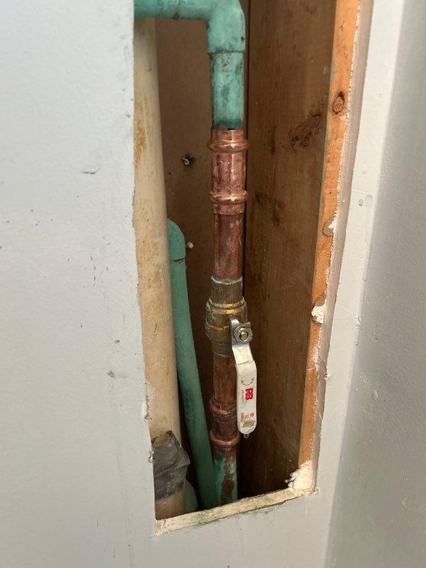 Responded to a pipe leak in a 20+ year old residential property. Upon inspection, accessed the affected plumbing through ceiling/wall cavity and identified a compromised copper water line showing significant age-related corrosion and oxidation. Removed drywall to expose the plumbing chase, revealing aged copper piping with darkened patina and PVC drain/vent lines with weathering and discoloration. Assessed the leak source at soldered joint connections where debris accumulation and pipe deterioration were evident. Installed ball valve isolation point on the copper supply line to control water flow to the affected section. Verified proper operation of shutoff valve and confirmed leak containment. Inspected surrounding wooden framing and adjacent green PVC piping for secondary water damage or structural concerns. The plumbing system displayed typical wear consistent with the home's age, with multiple material transitions between copper supply lines and plastic drainage components. Access 