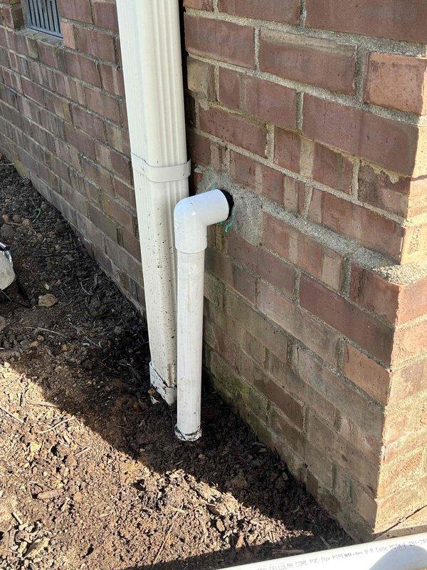 Responded to sump pump discharge concern where water was being released directly against the foundation. Upon arrival, assessed existing white PVC discharge system with elbow joint at foundation level that was directing water inadequately, causing moisture accumulation and soil saturation at the base of the 46-year-old brick structure. Installed pipe extension to the existing sump pump discharge line to redirect water flow further into the yard and away from the foundation. Excavated trench area as needed to accommodate new PVC sections and ensure proper grade for drainage. Connected extension piping to existing elbow fitting, securing all joints to prevent leaks and ensure long-term functionality. Verified water flow diverts appropriately away from foundation to prevent future moisture damage to lower brick courses and foundation integrity. Documented installation with photographs showing before and during work phases.