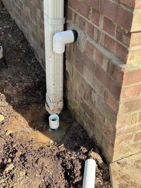 Responded to sump pump discharge concern where water was being released directly against the foundation. Upon arrival, assessed existing white PVC discharge system with elbow joint at foundation level that was directing water inadequately, causing moisture accumulation and soil saturation at the base of the 46-year-old brick structure. Installed pipe extension to the existing sump pump discharge line to redirect water flow further into the yard and away from the foundation. Excavated trench area as needed to accommodate new PVC sections and ensure proper grade for drainage. Connected extension piping to existing elbow fitting, securing all joints to prevent leaks and ensure long-term functionality. Verified water flow diverts appropriately away from foundation to prevent future moisture damage to lower brick courses and foundation integrity. Documented installation with photographs showing before and during work phases.