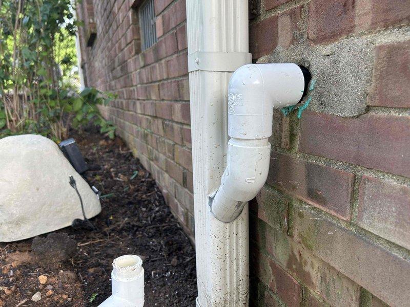 Assessed existing white PVC drainage pipe configuration attached to exterior brick wall that requires rerouting into the gutter system. The current setup consists of vertical and horizontal sections with elbow joints running along the foundation, separate from the aluminum downspout. Documented the weathered piping system and surrounding landscape conditions, including the planting bed and foundation area where modifications will be necessary. Will return to complete the rerouting work to properly integrate the drainage pipe with the existing gutter downspout system for improved water management.