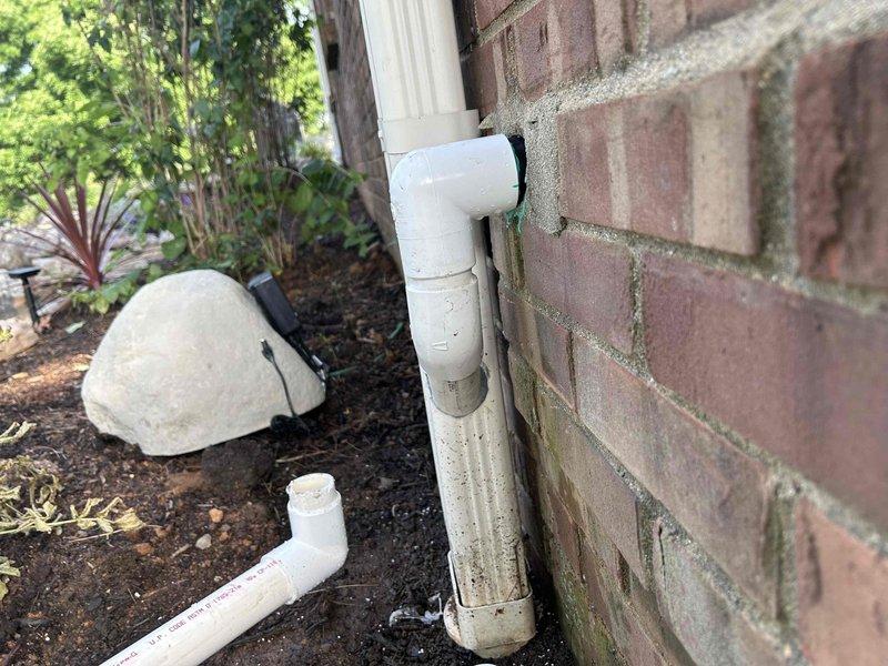 Assessed existing white PVC drainage pipe configuration attached to exterior brick wall that requires rerouting into the gutter system. The current setup consists of vertical and horizontal sections with elbow joints running along the foundation, separate from the aluminum downspout. Documented the weathered piping system and surrounding landscape conditions, including the planting bed and foundation area where modifications will be necessary. Will return to complete the rerouting work to properly integrate the drainage pipe with the existing gutter downspout system for improved water management.