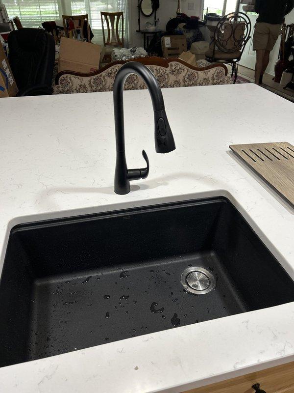 Installed new matte black pull-down kitchen faucet with gooseneck design on newly installed white quartz countertop with black composite undermount sink. Connected water supply lines to shut-off valves, installed stainless steel drain assembly with P-trap configuration using white PVC piping, and applied silicone sealant around faucet base and sink perimeter to ensure proper waterproofing.