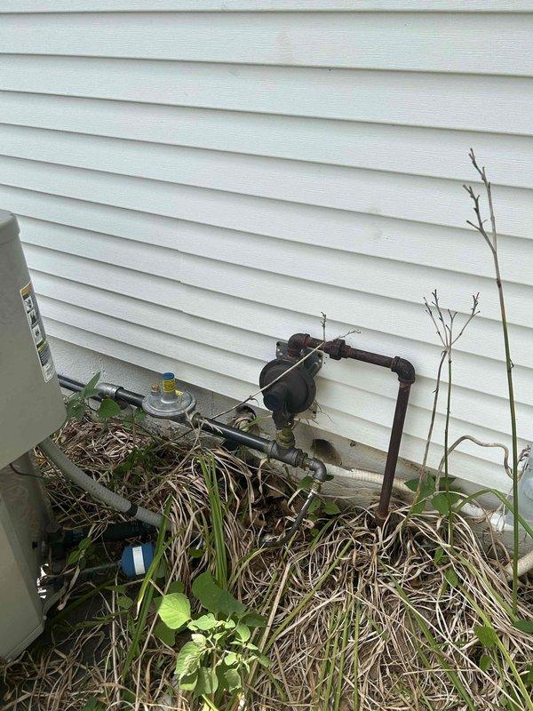 Completed generator installation with final connections and system verification. Documented exterior equipment placement including transfer switch, condenser unit, and associated utility connections mounted to building exterior. All electrical connections secured and tested for proper operation. Site conditions noted with vegetation overgrowth around equipment requiring future maintenance access considerations.