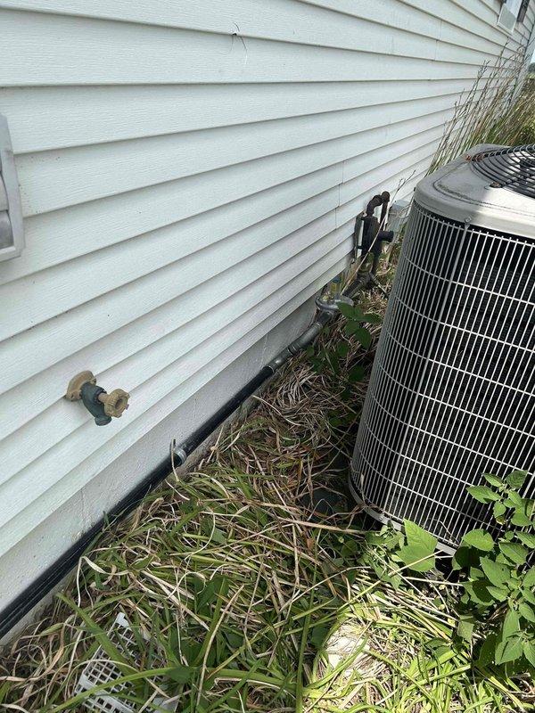 Completed generator installation with final connections and system verification. Documented exterior equipment placement including transfer switch, condenser unit, and associated utility connections mounted to building exterior. All electrical connections secured and tested for proper operation. Site conditions noted with vegetation overgrowth around equipment requiring future maintenance access considerations.