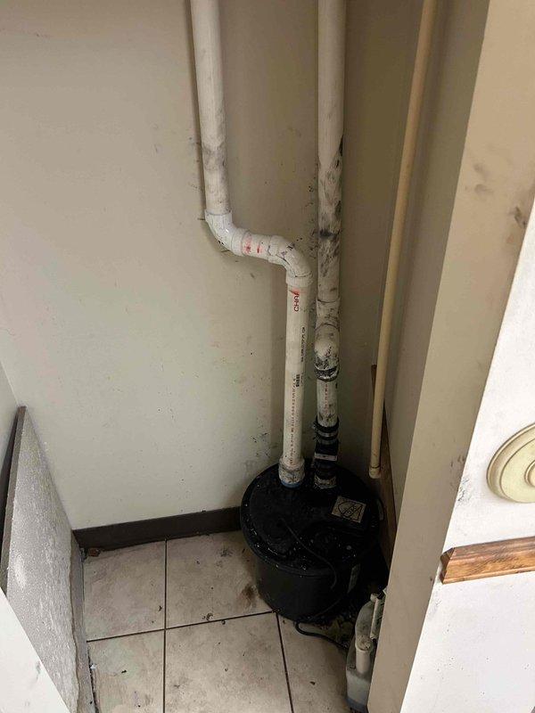 Removed and replaced existing sump pump system in utility room. Installed new black cylindrical sump pump unit at base of white PVC drainage piping configuration, ensuring proper connections to vertical discharge lines and establishing reliable water removal capability for the space.