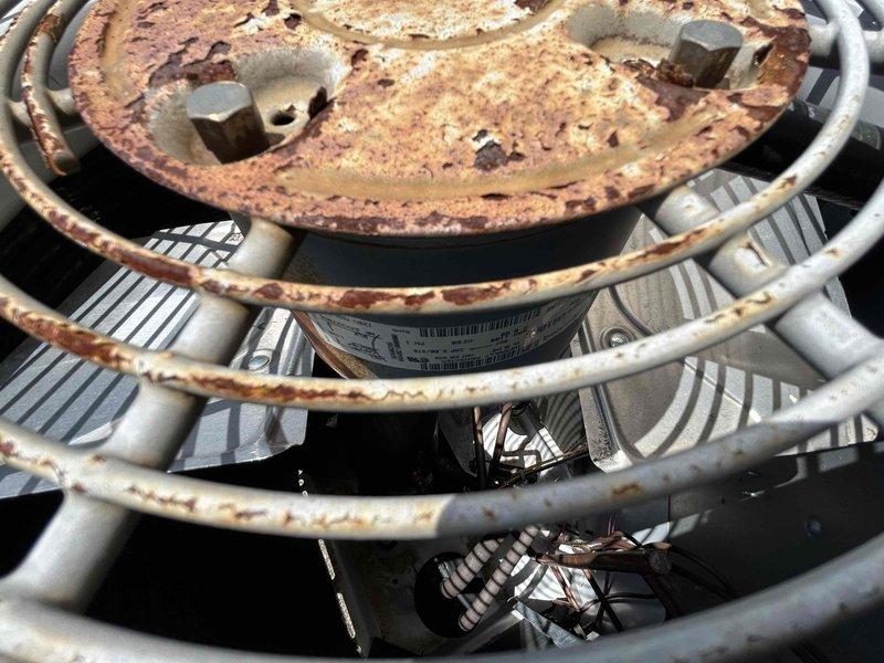 Responded to no cooling call on 8-9 year old electric HVAC system with reported popping sounds and breaker tripping. Upon arrival, performed visual inspection of outdoor condensing unit and discovered extensive rust and corrosion on condenser fan motor assembly and top protective grille. The circular metal plate above the fan motor showed heavy oxidation with significant brown and orange discoloration, indicating prolonged moisture exposure and deterioration. Condenser fan motor housing and protective cage components displayed weathering consistent with outdoor exposure. Documented equipment condition with photographs showing severity of rust damage to fan assembly and guard structure. The corrosion on critical components and electrical issues with breaker tripping indicate potential safety concerns and compromised system integrity requiring immediate attention.