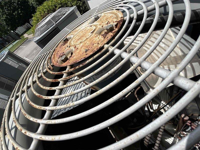 Responded to no cooling call on 8-9 year old electric HVAC system with reported popping sounds and breaker tripping. Upon arrival, performed visual inspection of outdoor condensing unit and discovered extensive rust and corrosion on condenser fan motor assembly and top protective grille. The circular metal plate above the fan motor showed heavy oxidation with significant brown and orange discoloration, indicating prolonged moisture exposure and deterioration. Condenser fan motor housing and protective cage components displayed weathering consistent with outdoor exposure. Documented equipment condition with photographs showing severity of rust damage to fan assembly and guard structure. The corrosion on critical components and electrical issues with breaker tripping indicate potential safety concerns and compromised system integrity requiring immediate attention.