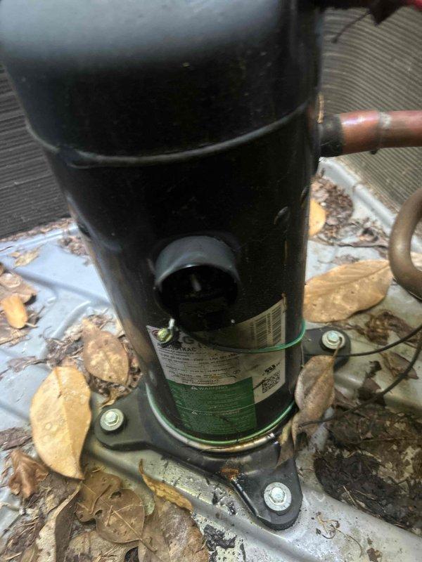 # HVAC Service Call - No Cooling Condition

Responded to service call for air conditioning system not providing cooling. Unit is approximately 3 years old. Upon arrival, performed visual inspection of outdoor condensing unit and found compressor with significant debris accumulation around base and mounting surface.

Inspected compressor unit and refrigerant line connections. Noted compressor mounted on metal base plate with copper refrigerant lines connected. Observed deterioration of mounting surface with peeling paint and rust-colored staining present. Heavy accumulation of dried leaves and organic debris found throughout equipment area and on base platform.

Cleaned debris from around compressor unit and base plate to ensure proper airflow and prevent future operational issues. Verified all electrical connections secure. Checked refrigerant line connections for proper sealing and integrity. Compressor unit itself appeared intact with manufacturer labeling visible and all connections