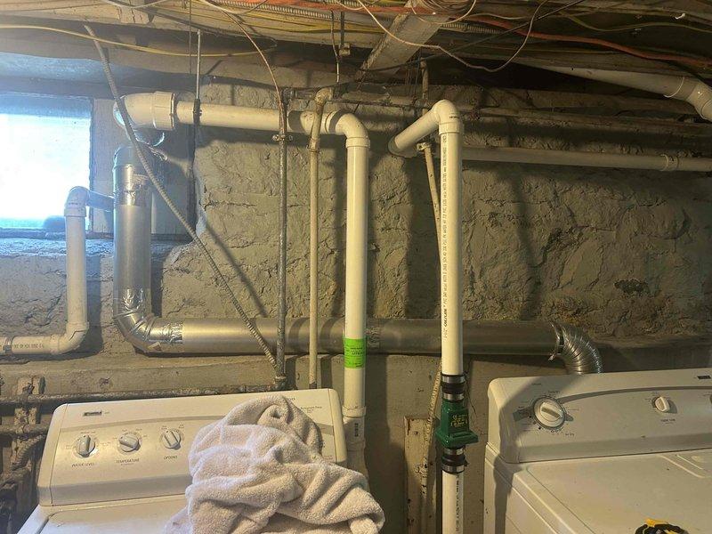 Conducted on-site assessment of basement laundry area to provide estimate for relocating dryer vent. Evaluated existing utility infrastructure including plumbing lines, electrical conduits, and current laundry equipment configuration to determine optimal routing path for dryer exhaust. Documented existing conditions with photos showing washing machine placement, PVC drainage system, water filtration equipment, exposed ceiling joists, and concrete block wall structure to develop relocation plan and material requirements for future dryer vent installation.