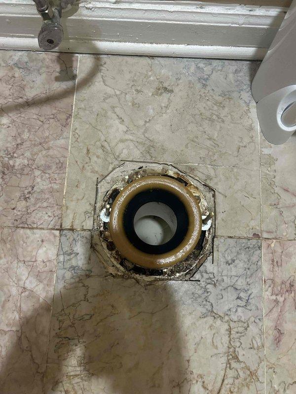 Responded to service call for unstable toilet that was rocking and moving during use. Upon inspection, removed toilet to access the flange and discovered a deteriorated toilet flange with significant wear, staining, and compromised structural integrity. The existing flange showed brown discoloration and degradation around the mounting area, which was causing the toilet to lose its secure connection to the floor. Documented the condition of the damaged flange and surrounding tile flooring for repair assessment.