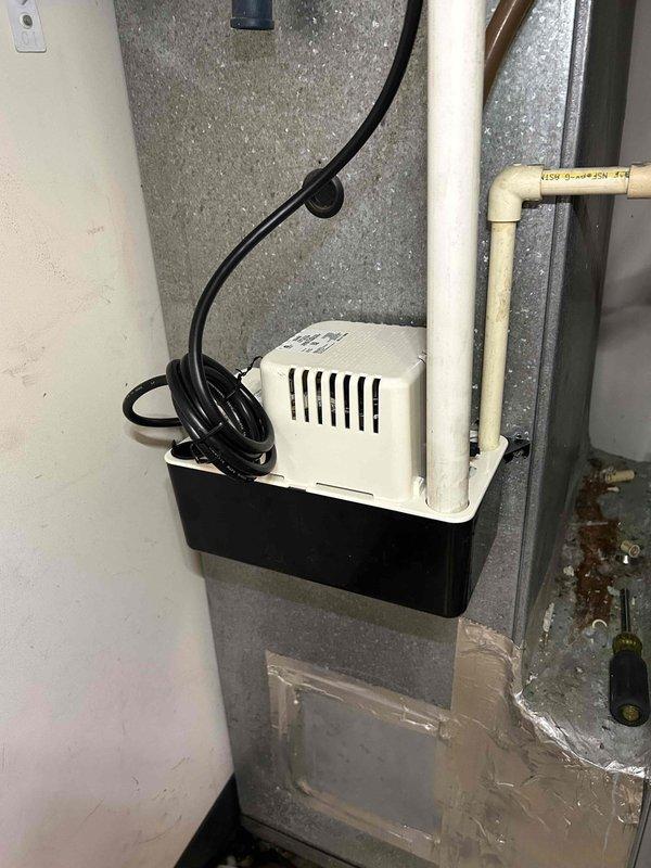 Responded to water leak complaint at indoor HVAC unit in utility room that began the previous night. Upon inspection, found condensate pump system with drain pan positioned beneath Carrier furnace air handler showing signs of moisture accumulation on metallic surfaces. Documented existing condensate management setup including white plastic condensate pump unit on black drain pan with connected PVC drainage piping and verified electrical connections to wall outlet.