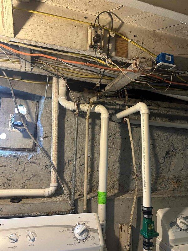Removed existing discharge and vent piping configuration and rerouted lines through the bandboard in the basement. Cut out old PVC sections and installed new white PVC piping with appropriate elbows and joints, running the system from the interior basement wall through the foundation to the exterior of the brick structure. Completed installation includes proper venting and drainage routing with secure mounting and connections throughout the basement utility area.