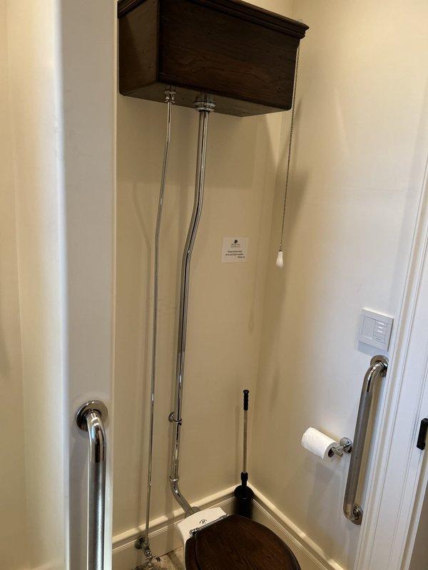 Responded to service call for multiple plumbing issues including a noisy toilet in the half bath, unsecured hydrant at barn wall, and leak under master bathroom. Inspected vintage high-tank toilet system with overhead wooden tank and pull-chain flush mechanism; diagnosed intermittent running noise and assessed flush valve and fill valve operation. Examined water supply shutoff valves showing corrosion and oxidation on copper piping with evidence of moisture damage and water staining on surrounding wall surface, indicating active or previous leak condition. Secured hydrant mounting at barn location and documented all findings with photographic evidence for repair recommendations.