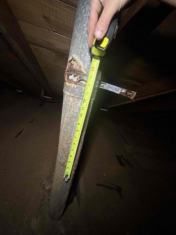 Responded to customer report of rusted furnace flue pipe with small hole. Upon inspection in confined attic space, located vertical galvanized metal vent pipe with significant rust deterioration on metal connector clamp joining two duct sections. Documented extensive corrosion with reddish-brown oxidation on clamp and surface staining on lower duct section. Measured affected ductwork and photographed rust damage for repair assessment.