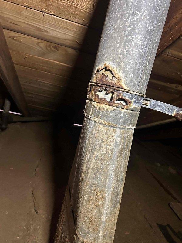 Responded to customer report of rusted furnace flue pipe with small hole. Upon inspection in confined attic space, located vertical galvanized metal vent pipe with significant rust deterioration on metal connector clamp joining two duct sections. Documented extensive corrosion with reddish-brown oxidation on clamp and surface staining on lower duct section. Measured affected ductwork and photographed rust damage for repair assessment.