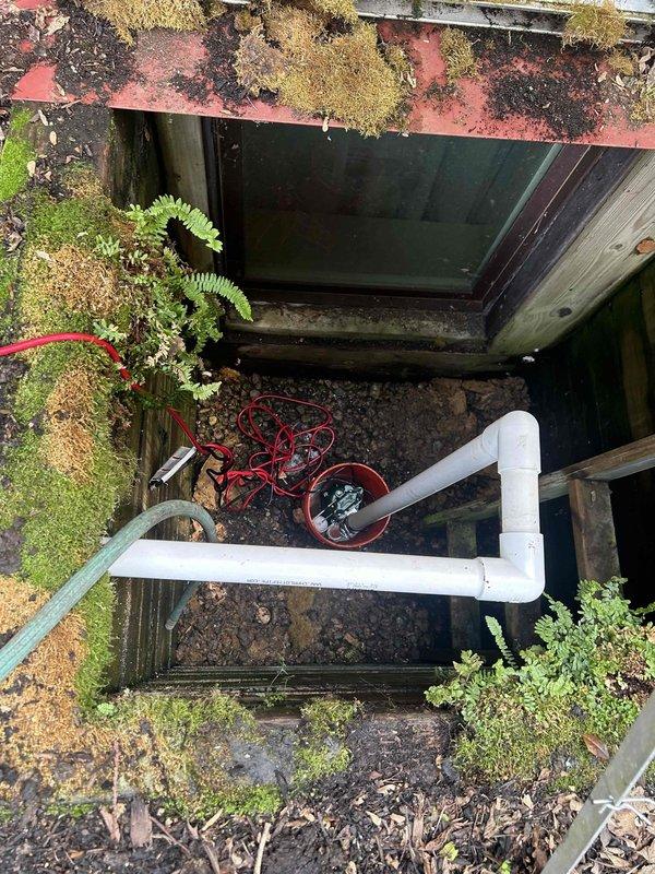 Installed new outdoor sump pump system to address flooding issues at basement window well. Excavated 6-8 inches below grade and set pump basin with sump pump unit. Ran PVC discharge line vertically through wooden window well structure and terminated into existing gutter system with proper air gap to prevent backflow. System tested and operational upon completion.