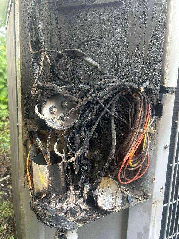 Responded to no cooling complaint on Puron system. Upon arrival, discovered catastrophic electrical failure in outdoor unit's control compartment with extensive fire damage. Contactor, capacitor, and control wiring sustained severe heat damage with melted insulation, charred components, and fused connections. External evidence of electrical fire visible with significant soot and burn marks on adjacent wall above unit. All damaged electrical components including contactor, capacitor, and control wiring harness require complete replacement. Compartment interior shows heat discoloration and corrosion throughout. System remains inoperable and unsafe for operation until full electrical component replacement completed. Documented extensive damage with photos for reference.