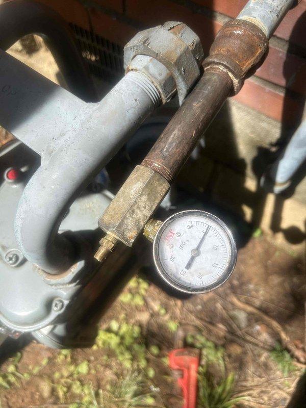 Responded to vacant property for gas leak pressure test following previous technician's assessment. Connected pressure gauge to exterior gas line system to conduct pressurization test on weathered piping infrastructure showing significant age and corrosion. Performed pressure testing procedures on gas distribution system to identify potential leak points, with gauge monitoring maintaining zero PSI reading during inspection. Documented condition of aging metal pipes, corroded fittings, and deteriorated white-painted elbow joints requiring evaluation for system integrity and safety compliance.