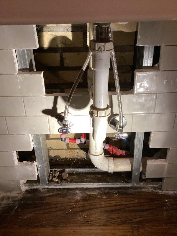 Accessed concealed plumbing leak by cutting through drywall and tile at bathroom vanity wall cavity. Located leak source at supply line connection point behind sink installation. Repaired compromised fitting and replaced damaged braided stainless steel supply line. Reassembled white PVC drain assembly including P-trap configuration with proper slip-joint connections. Installed new shut-off valves and verified secure connections on both hot and cold water supply lines. Tested all plumbing connections under pressure to confirm leak elimination and proper drainage function. System verified operational with no active leaks detected prior to departure.