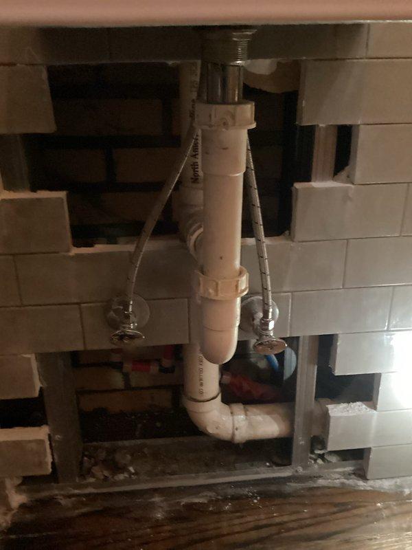 Accessed concealed plumbing leak by cutting through drywall and tile at bathroom vanity wall cavity. Located leak source at supply line connection point behind sink installation. Repaired compromised fitting and replaced damaged braided stainless steel supply line. Reassembled white PVC drain assembly including P-trap configuration with proper slip-joint connections. Installed new shut-off valves and verified secure connections on both hot and cold water supply lines. Tested all plumbing connections under pressure to confirm leak elimination and proper drainage function. System verified operational with no active leaks detected prior to departure.