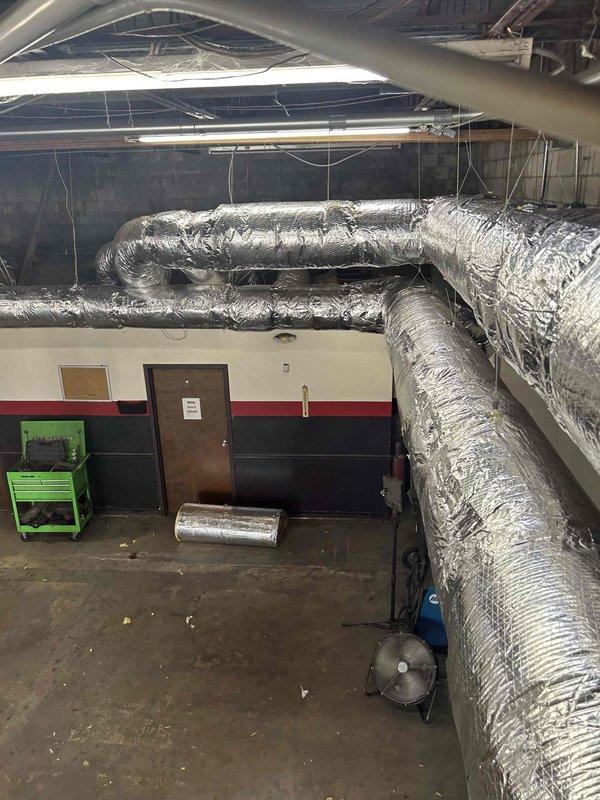 Wrapped spiral ductwork system in tire shop facility with R8 foilback insulation on both supply and return lines to eliminate condensation and leaking caused by high humidity environment contacting cold ductwork during system operation. All seams were sealed with foil tape to prevent temperature loss and ensure proper vapor barrier. Additionally, installed auxiliary drain pan drain line using 3/4" pipe with four 3/4" 90-degree elbows, one 1-1/2" 90-degree elbow, one 1-1/2" bushing to 3/4", and 1-1/2" pipe section to properly route condensate drainage.