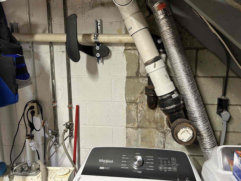 Conducted on-site assessment of existing sump pump discharge system in basement utility area to provide relocation estimate. Evaluated current PVC discharge piping configuration, including horizontal runs along ceiling joists and wall-mounted sections. Inspected mechanical room conditions, noting moisture staining on concrete block walls and existing plumbing infrastructure including copper piping, flexible HVAC ductwork, and electrical systems. Documented exposed ceiling joists, structural framing, and routing pathways for potential discharge line relocation. Photographed current installation to support estimate preparation for municipal discharge system modifications.