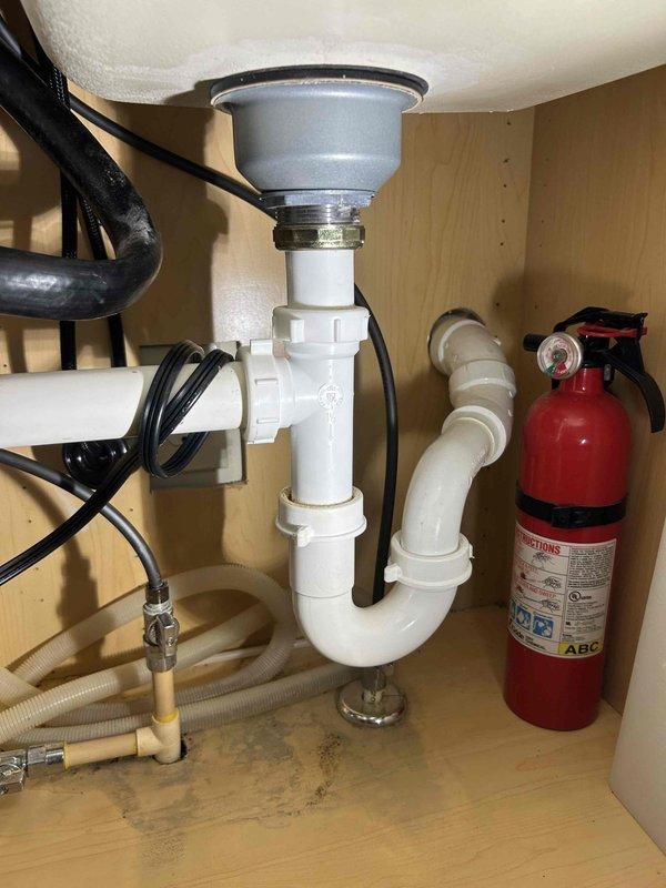 Responded to report of intermittent water leak under kitchen sink. Upon inspection, examined garbage disposal unit, P-trap assembly, and dishwasher connection points. Found evidence of minor water accumulation on cabinet floor beneath plumbing components. Inspected all compression fittings on white PVC P-trap, garbage disposal mounting flange, discharge connections, and dishwasher drain line. Verified integrity of all visible plumbing connections and identified potential source areas requiring monitoring for future leakage.