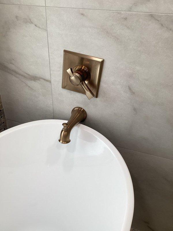 Provided plumbing estimate for replacement of existing tub filler fixture in historic two-story residence built in 1880. Current wall-mounted tub spout and shower valve control feature brushed bronze finish that was installed during previous renovation but does not match other bathroom fixtures. Assessed installation complexity due to fixture mounting behind marble-look tile wall and documented existing configuration including square escutcheon plate with lever handle and wall-mounted spout assembly. Customer expressed concerns regarding proper removal and reinstallation to avoid tile damage during replacement process.