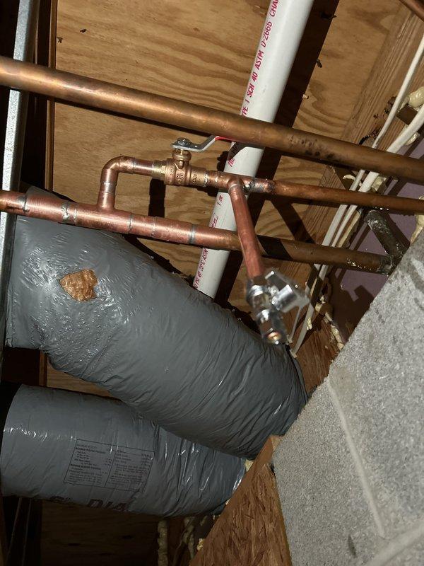 Returned to site for concrete pour as scheduled. Documented completed water heater installation in basement utility area, showing insulated tank with copper supply and distribution piping, brass valves, and proper connections to building systems. Photographed exposed plumbing configuration in attic space, including copper pipe U-bend trap assembly mounted to structural joists. Noted visible corrosion and oxidation on copper surfaces with blue-green patina formation and surface deterioration requiring future monitoring. All rough plumbing work verified and ready for concrete floor installation to proceed.