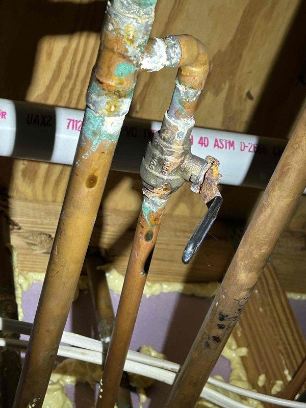 Returned to site for concrete pour as scheduled. Documented completed water heater installation in basement utility area, showing insulated tank with copper supply and distribution piping, brass valves, and proper connections to building systems. Photographed exposed plumbing configuration in attic space, including copper pipe U-bend trap assembly mounted to structural joists. Noted visible corrosion and oxidation on copper surfaces with blue-green patina formation and surface deterioration requiring future monitoring. All rough plumbing work verified and ready for concrete floor installation to proceed.