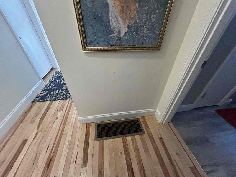 Returned to property for follow-up inspection of HVAC installation with focus on hardwood flooring condition near register locations. Documented wall-mounted vent installation in wood-paneled area and floor register placement in hardwood entryway. Photographed both installations to verify proper integration with finished surfaces and confirm no damage occurred to wood flooring or paneling during system installation.