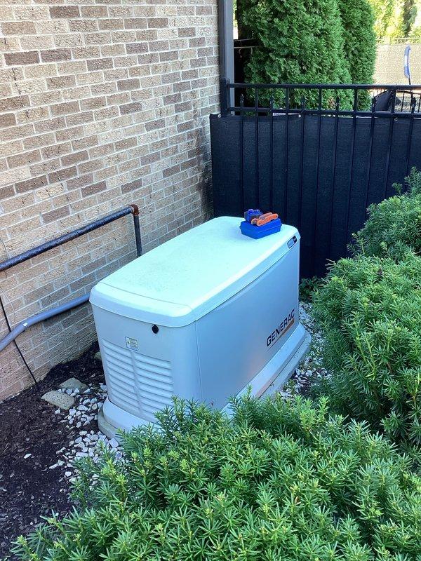 Completed on-site assessment of existing Generac standby generator installation to provide estimate for repositioning unit. Generator currently located on white gravel pad against exterior brick wall with adequate clearance and drainage. Unit features proper ventilation and professional mounting on stable base. Documented current installation configuration including gas supply line routing, electrical conduit placement, and proximity to structure. Evaluated existing infrastructure including corroded gas piping with visible rust requiring replacement during relocation. Assessed access constraints in narrow side yard area with moss-covered pavers indicating drainage considerations for new placement. Photographed current setup showing generator positioning relative to privacy fencing, landscaping, and building exterior. Documented conduit runs and utility connections for planning relocation scope. Identified need to address deteriorated gas line and mounting hardware as part of reposition