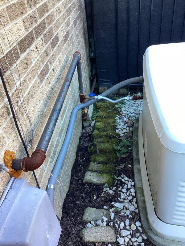 Completed on-site assessment of existing Generac standby generator installation to provide estimate for repositioning unit. Generator currently located on white gravel pad against exterior brick wall with adequate clearance and drainage. Unit features proper ventilation and professional mounting on stable base. Documented current installation configuration including gas supply line routing, electrical conduit placement, and proximity to structure. Evaluated existing infrastructure including corroded gas piping with visible rust requiring replacement during relocation. Assessed access constraints in narrow side yard area with moss-covered pavers indicating drainage considerations for new placement. Photographed current setup showing generator positioning relative to privacy fencing, landscaping, and building exterior. Documented conduit runs and utility connections for planning relocation scope. Identified need to address deteriorated gas line and mounting hardware as part of reposition