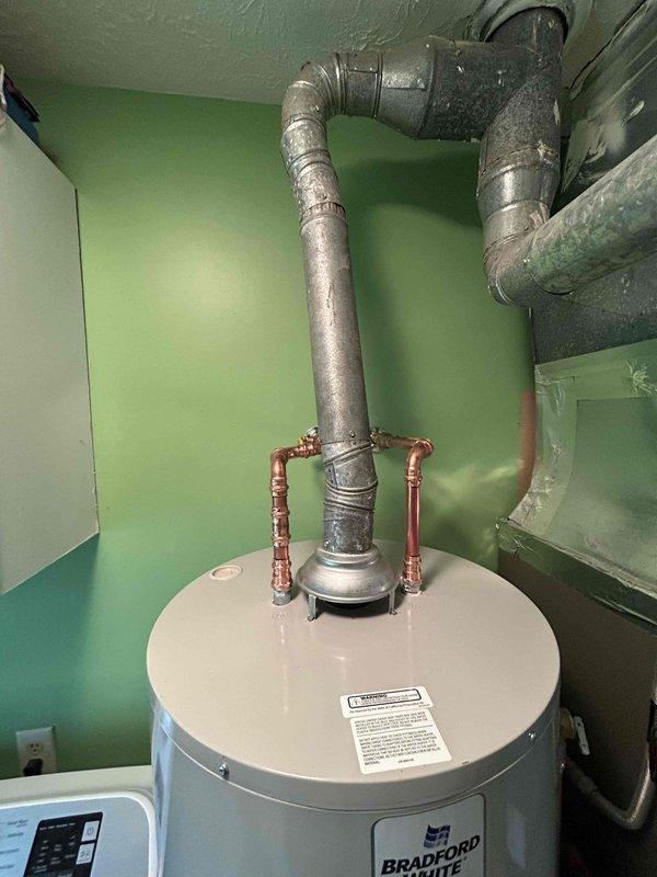 Responded to reported water heater leak with potential ceiling damage. Upon inspection, identified active leak from Bradford White water heater with compromised copper pipe connections at top of unit. Observed water staining on utility room floor tiles indicating ongoing moisture intrusion. Assessed plumbing connections, compression fittings, and vent ductwork for integrity. Documented existing conditions including scattered insulation materials and water damage patterns. Photographed unit configuration, piping layout, and affected areas for repair planning and customer records.