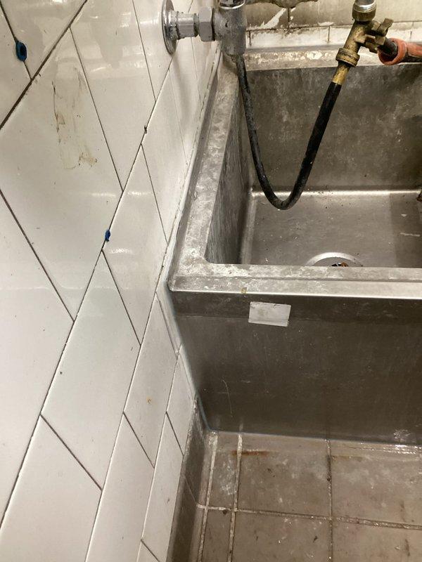 Responded to commercial water leak at mop sink location. Upon inspection, identified active leak in P-trap assembly beneath industrial stainless steel utility sink. Removed existing mop sink unit to access plumbing connections and diagnose source of water intrusion. Inspected white PVC drain configuration and associated trap assembly in crawlspace area. Repaired defective drain connections, replaced worn P-trap components, and verified proper seal on all plumbing fittings. Reinstalled utility sink with chrome wall-mounted faucet and utility hose connections. Tested system for leaks and confirmed proper drainage function. All plumbing connections secured and operational.