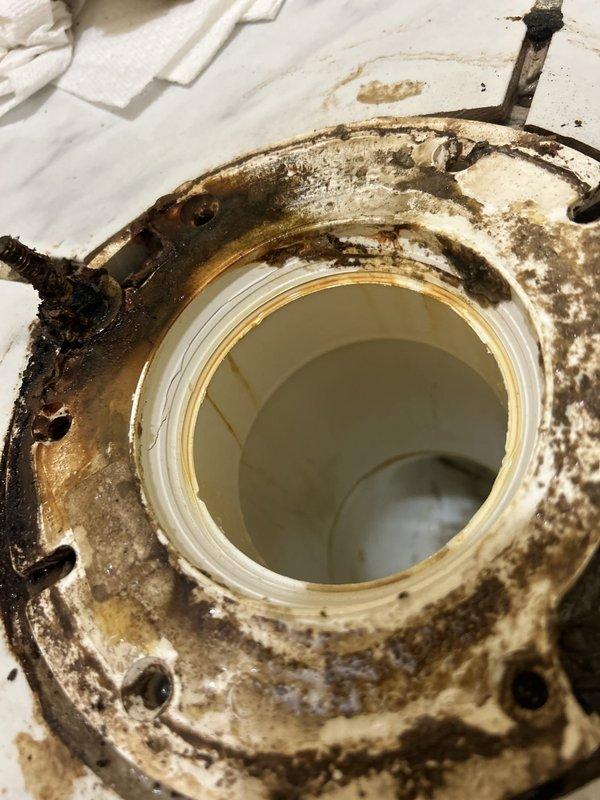 Responded to water leak originating from master bathroom toilet affecting adjacent master bedroom closet carpeting. Upon inspection, discovered severely corroded toilet flange with extensive rust damage and deterioration of mounting bolts. The metal flange exhibited significant oxidation from prolonged moisture exposure, compromising the seal between the toilet and drain pipe. Removed toilet to access and assess the failed flange assembly. Documented the extensive corrosion on both the metal flange components and mounting hardware, which had allowed water to seep beneath the fixture and migrate through the wall into the adjacent closet space. The white PVC pipe connection remained intact, but the surrounding metal mounting flange required complete replacement due to structural failure from rust deterioration.