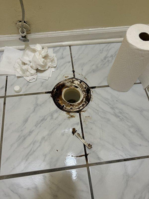 Responded to water leak originating from master bathroom toilet affecting adjacent master bedroom closet carpeting. Upon inspection, discovered severely corroded toilet flange with extensive rust damage and deterioration of mounting bolts. The metal flange exhibited significant oxidation from prolonged moisture exposure, compromising the seal between the toilet and drain pipe. Removed toilet to access and assess the failed flange assembly. Documented the extensive corrosion on both the metal flange components and mounting hardware, which had allowed water to seep beneath the fixture and migrate through the wall into the adjacent closet space. The white PVC pipe connection remained intact, but the surrounding metal mounting flange required complete replacement due to structural failure from rust deterioration.