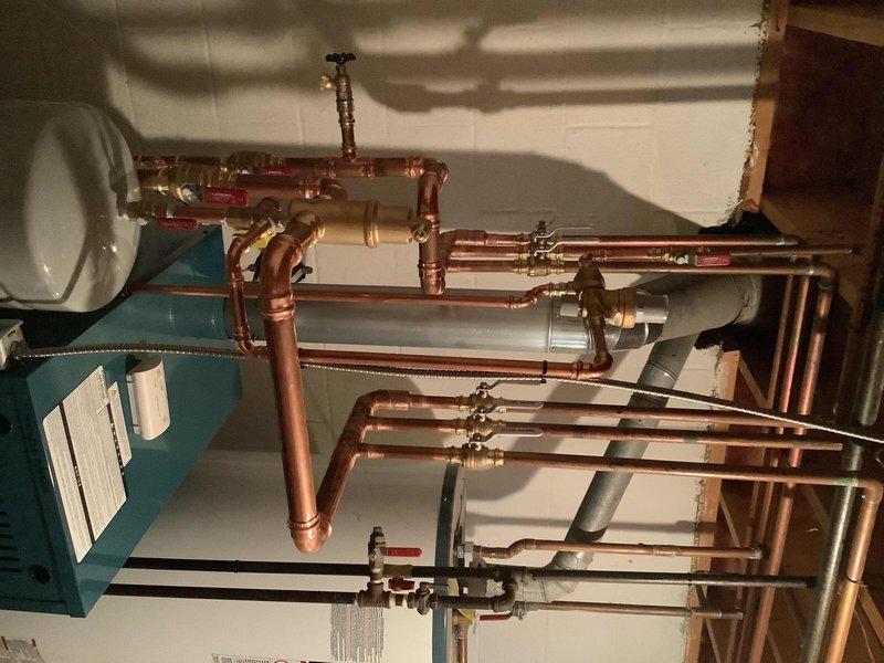 Completed boiler installation with comprehensive piping system setup in basement utility area. Installed new teal/blue boiler unit with associated copper piping network, valves, and fittings, ensuring proper integration with existing water heater. All components properly mounted, connected, and verified operational per installation specifications.
