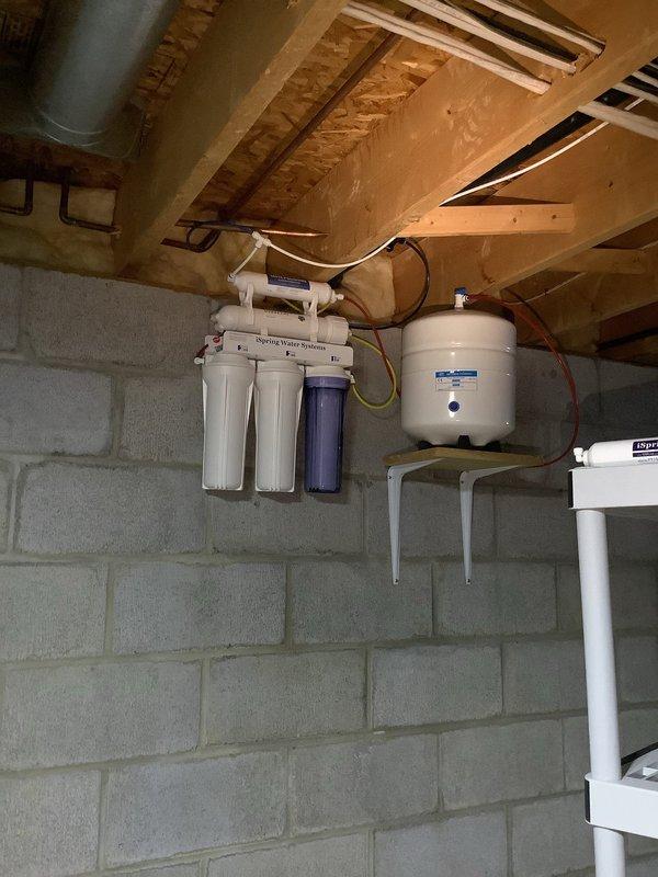 Completed inspection of residence for installation of whole home reverse osmosis system and dedicated drinking water filtration. Assessed current kitchen setup featuring dual faucet configuration with existing drinking water dispenser. Evaluated basement utility area which currently houses a Spring Water Systems filtration unit with multi-stage filtration (two white housings, one blue) and pressure tank mounted on concrete block wall.